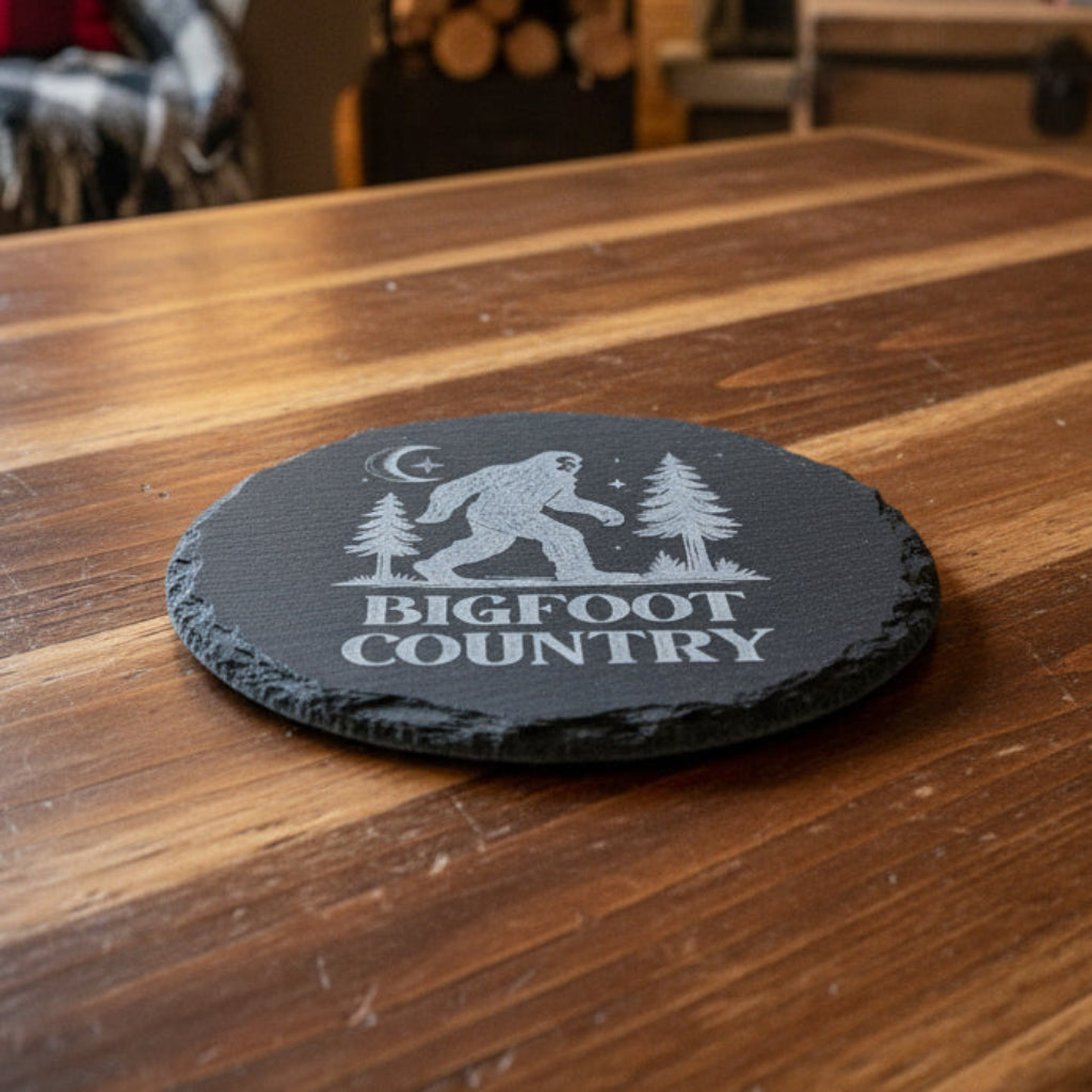 Black round slate with 'Bigfoot Country' design featuring a Sasquatch, trees, and moon.