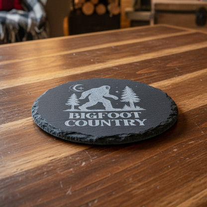 Black round slate with 'Bigfoot Country' design featuring a Sasquatch, trees, and moon.