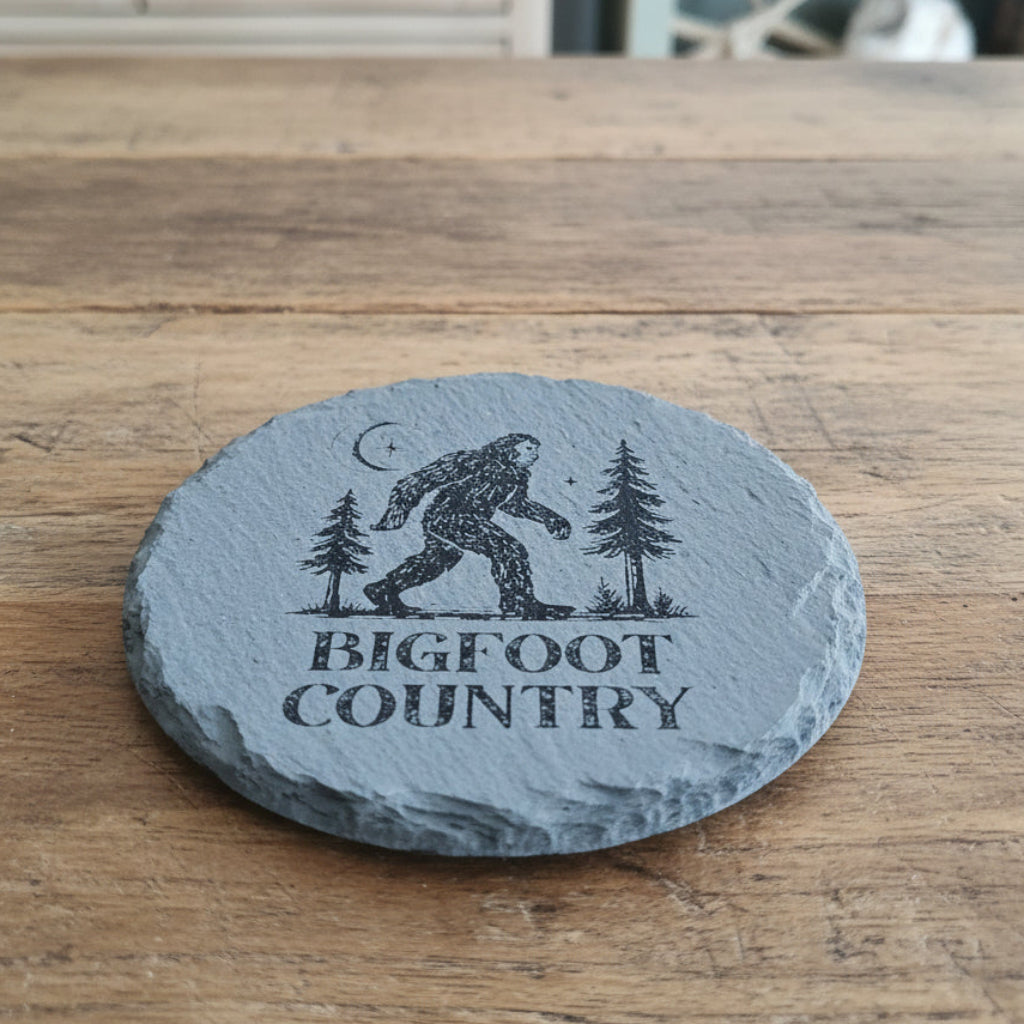 Round slate coaster with Bigfoot design and 'Bigfoot Country' text on a woven basket background.