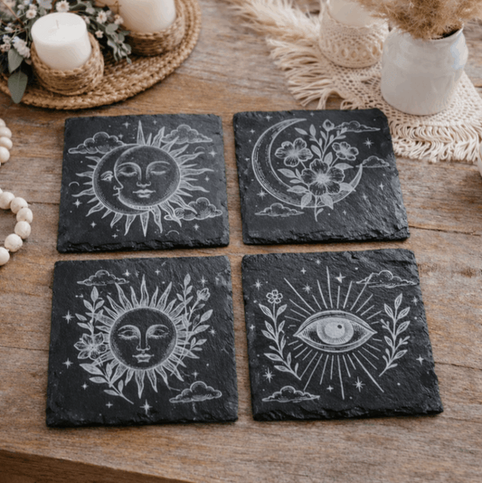 Four black slate coasters with boho celestial designs on a wooden surface.