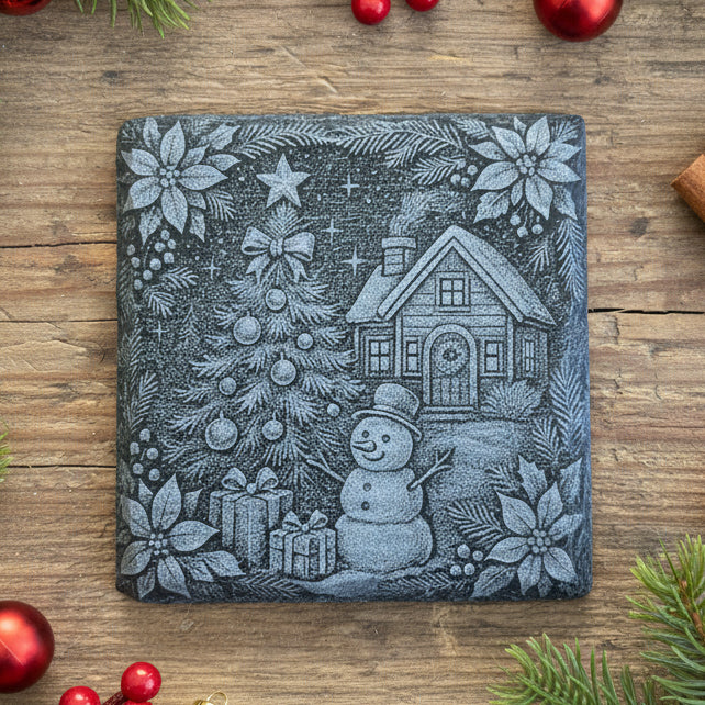 Decorative slate stone coaster with a Christmas scene featuring a house, tree, snowman, and presents on a wooden surface.