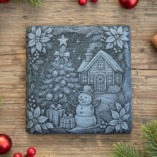 Decorative slate stone coaster with a Christmas scene featuring a house, tree, snowman, and presents on a wooden surface.