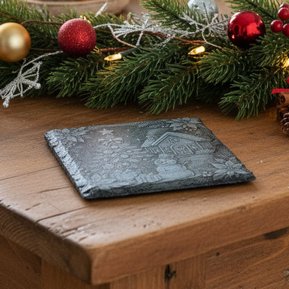 Decorative stone with a Christmas scene featuring a house, tree, snowman, and presents on a wooden surface.