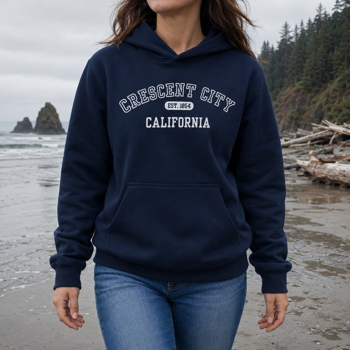 A woman wearing a navy blue hoodie with 'CRESCENT CITY CALIFORNIA' printed in white on the chest, standing on a beach with ocean waves and forest in the background.