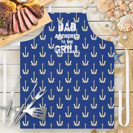 Blue Apron with white straps with white anchors and says Dad Anchored to the Grill with utensils in the font.