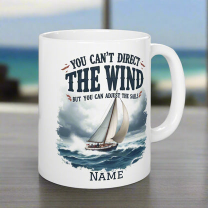 A white 11 oz. mug with a sailboat on the ocean with text You Can't Direct the Wind But You Can Adjust the Sails with personalized name.