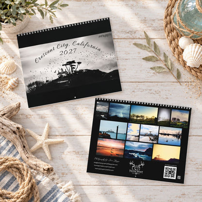 Front and back spiral-bound 2027 wall calendar  displayed on a rustic white wood surface with nautical props. The top calendar shows a black-and-white Crescent City, California, 2027 cover with a lighthouse silhouette and back shows all month pictures of Northern California Coast.