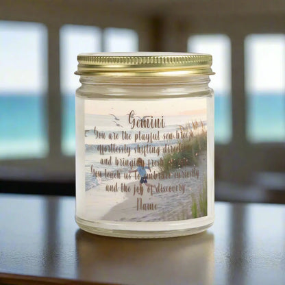 Elegant Gemini Zodiac hand-poured coconut apricot wax scented candle in a 9 oz clear glass jar with gold lid, featuring a scene of crashing waves with a man in the distance on a rock with the bright sun shining off of the waves girl running  on the beach to the ocean water and an inspirational quote personalized. A beach-inspired astrology gift, perfect for relaxation and decor.