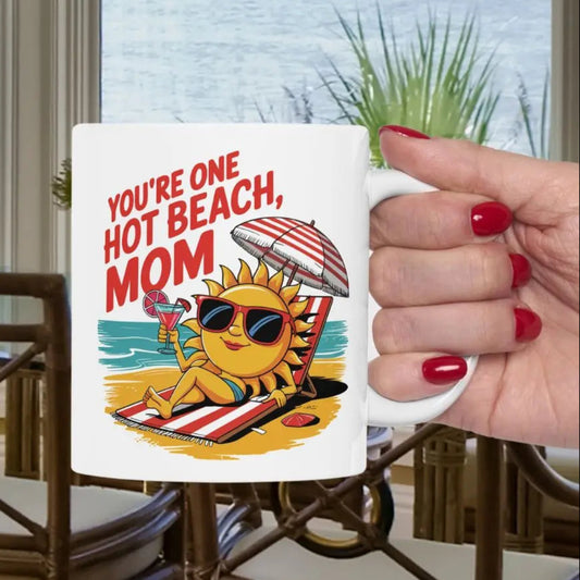 A white 11 oz mug with a sexy sun laying on a beach towel holding a drink on the beach that says You're One Hot Beach Mom.