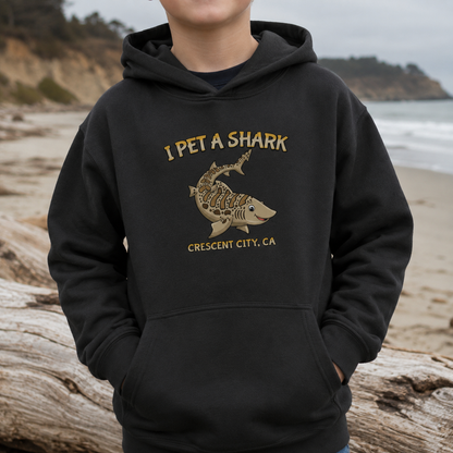 A person wearing a black hoodie on a beach, with the hoodie featuring a gold graphic of a shark skeleton and the text 'I PET A SHARK' and 'CRESCENT CITY, CA'.