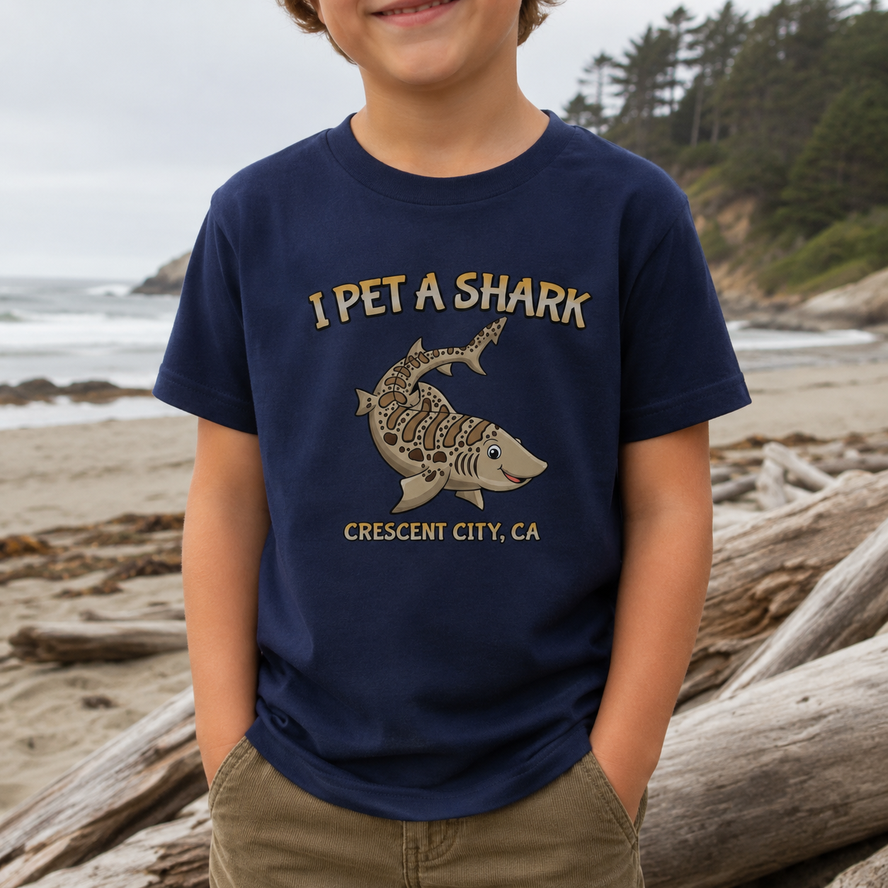 A child wearing a navy blue t-shirt with a graphic that reads 'I PET A SHARK' and 'CRESCENT CITY, CA' with a shark illustration, standing on a beach with driftwood and trees in the background.