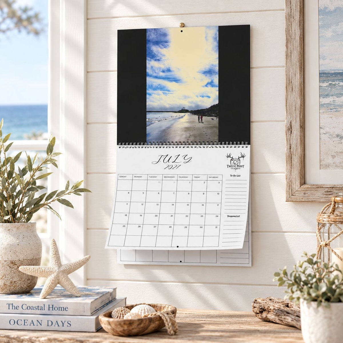 A wall-mounted July 2027 wall calendar monthly planning grid with to-do list and shopping list featuring Twelve Point Pics photography.