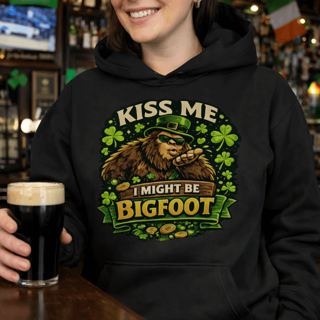Person wearing a black hoodie with Bigfoot blowing a kiss, surrounded by shamrocks and gold coins, wearing a leprechaun hat, with the text “Kiss Me, I Might Be Bigfoot”
