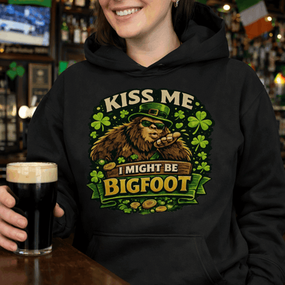 Person wearing a black hoodie with Bigfoot blowing a kiss, surrounded by shamrocks and gold coins, wearing a leprechaun hat, with the text “Kiss Me, I Might Be Bigfoot”