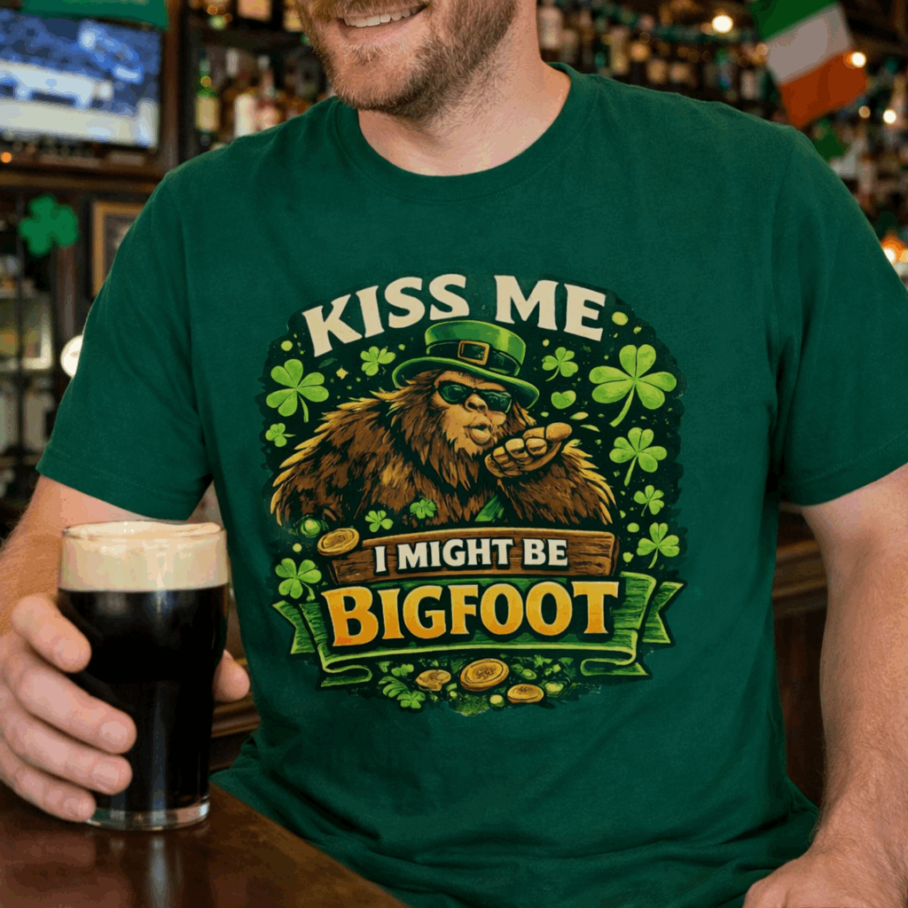 Person wearing a green t-shirt with Bigfoot illustration wearing a leprechaun hat, surrounded by shamrocks and gold coins, with the text “Kiss Me, I Might Be Bigfoot”.