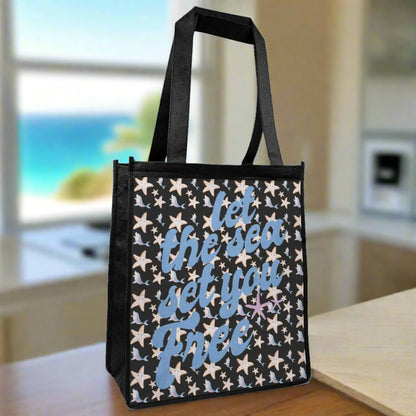 A black tote bag with cartoon starfish and dolphins in blue and pinks with blue fancy text that says let the sea set you free