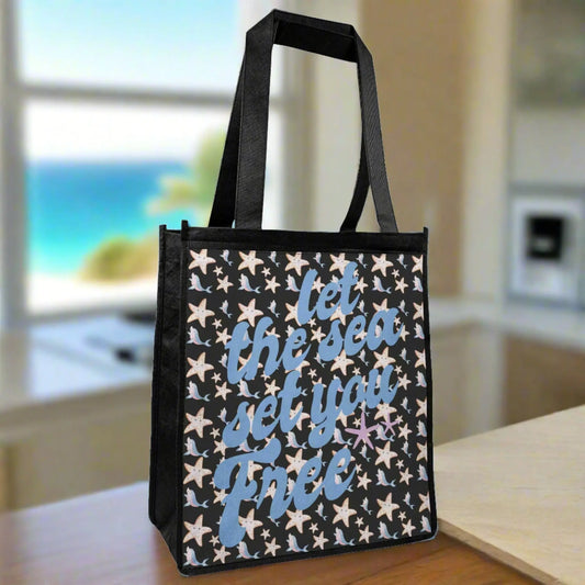 A black tote bag with cartoon starfish and dolphins in blue and pinks with blue fancy text that says let the sea set you free