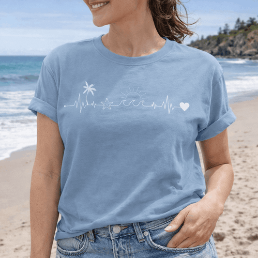 Woman wearing light blue t-shirt with a heartline that has a palm tree, sand dollar, sun with waves and a heart at the end.