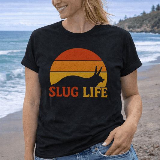 Person wearing a black t-shirt with 'Slug Life' retro graphic on a beach.