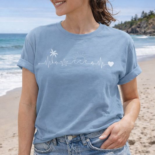 Woman wearing light blue t-shirt with a heartline that has a palm tree, sand dollar, sun with waves and a heart at the end.