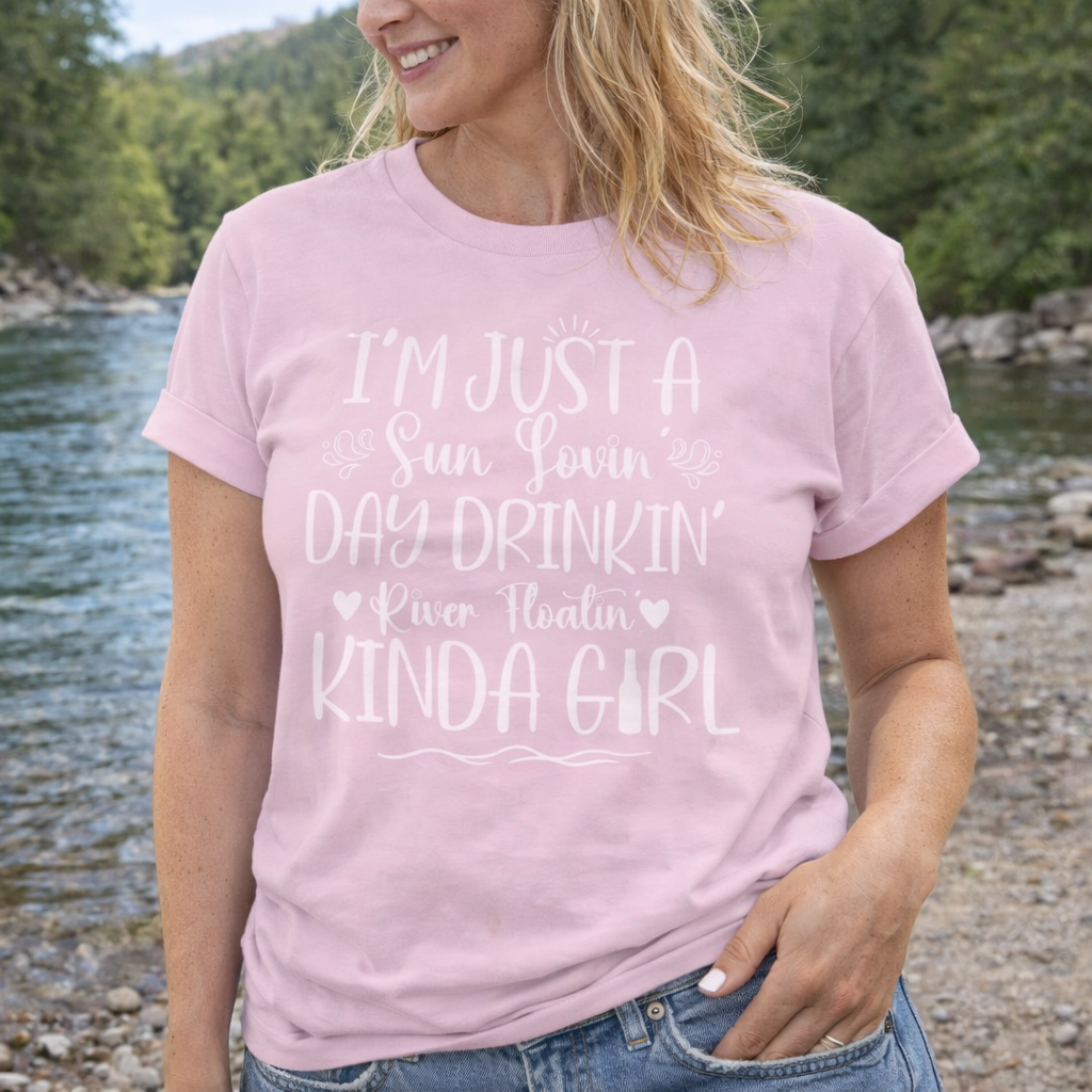 A woman wearing a pink t-shirt with the phrase "Sun-loving, Day-drinking, River-floating Kind of Girl" printed on the front with embellishments, featuring a relaxed fit and soft cotton fabric.
