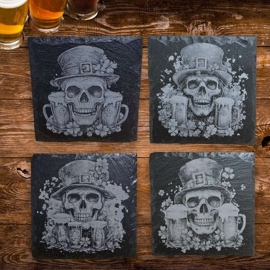 Four black slate coasters with skull and beer mug designs on a wooden surface.