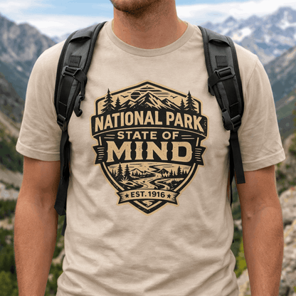 Person wearing a outdoor t-shirt with vintage badge graphic reading “National Park State of Mind” and “Est. 1916,” featuring mountains, trees, and a winding river.
