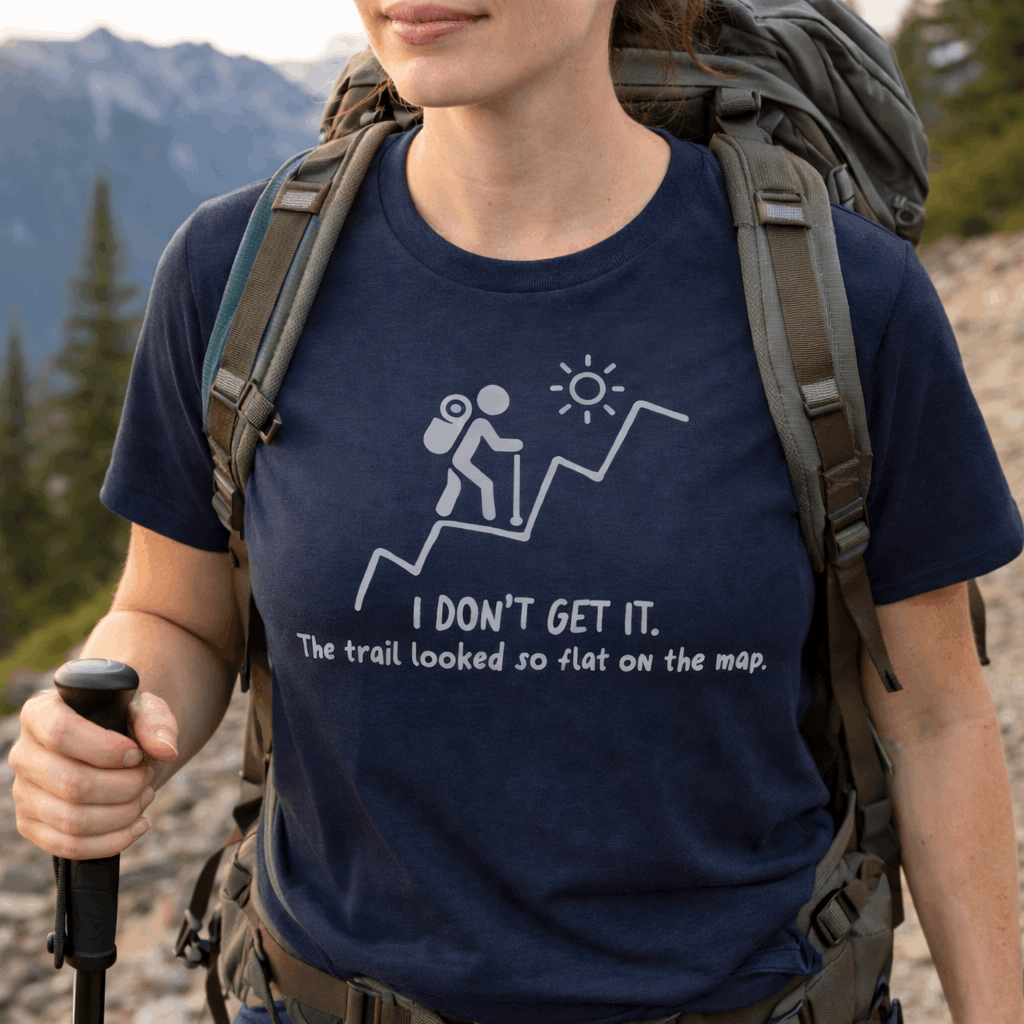 Person hiking with a blue hiking t-shirt with white graphic of a backpacker climbing a steep trail and text: “I DON’T GET IT. The trail looked so flat on the map.”