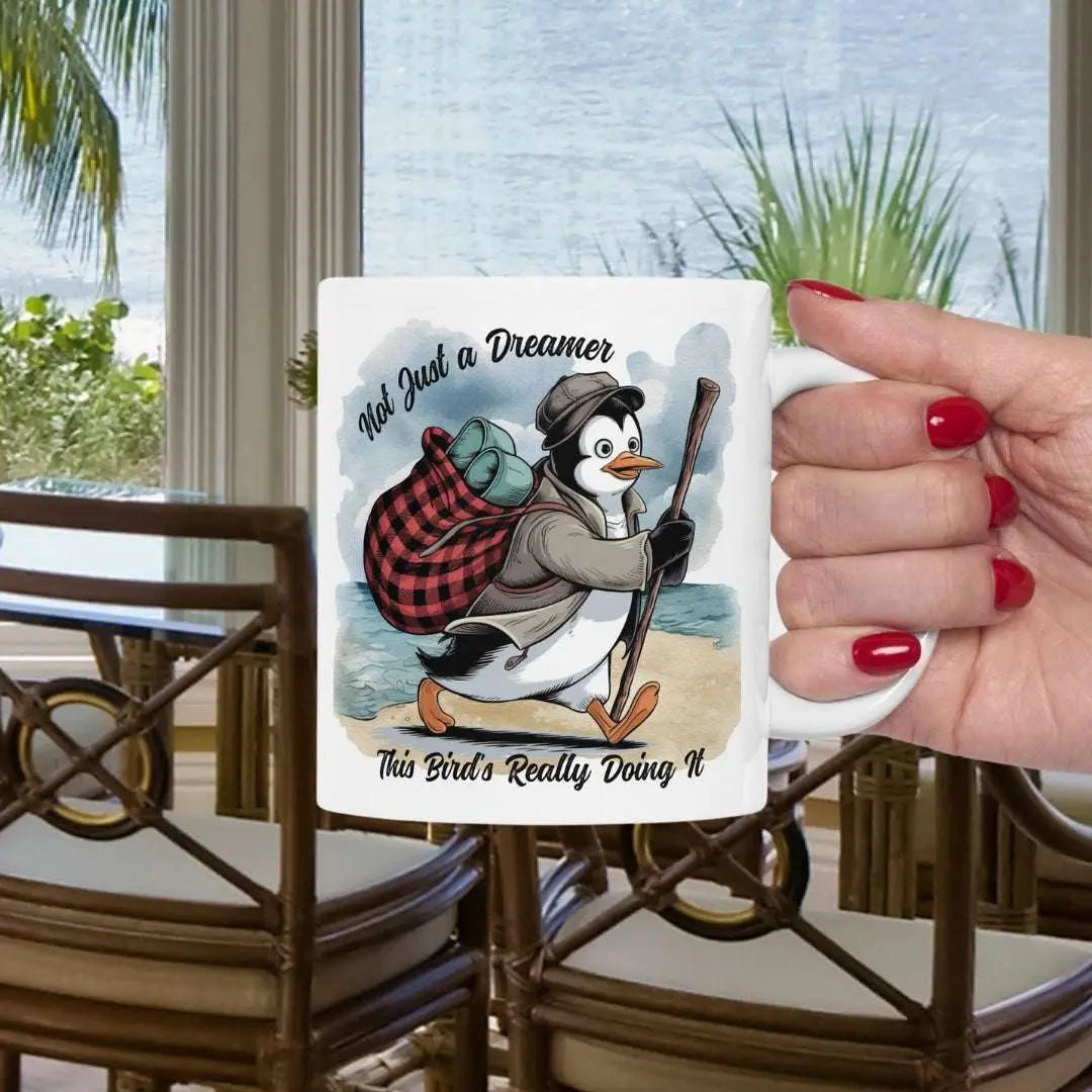 White 11 oz coffee mug featuring a fun beach design of a penguin running away on the sand with the text 'Not Just a Dreamer, This Bird’s Really Doing It.' A playful and unique gift for beach lovers and dreamers.