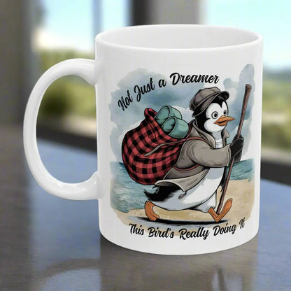 White 11 oz coffee mug featuring a fun beach design of a penguin running away on the sand with the text 'Not Just a Dreamer, This Bird’s Really Doing It.' A playful and unique gift for beach lovers and dreamers.