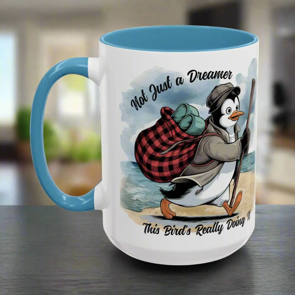 White 15 oz coffee mug with light blue interior and handle featuring a fun beach design of a penguin running away on the sand with the text 'Not Just a Dreamer, This Bird’s Really Doing It.' A playful and unique gift for beach lovers and dreamers.