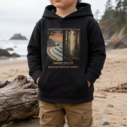 A child wearing a black Crescent City toddler hoodie featuring vintage redwood forest and Pacific coast Where the Redwoods Meet the Sea artwork.