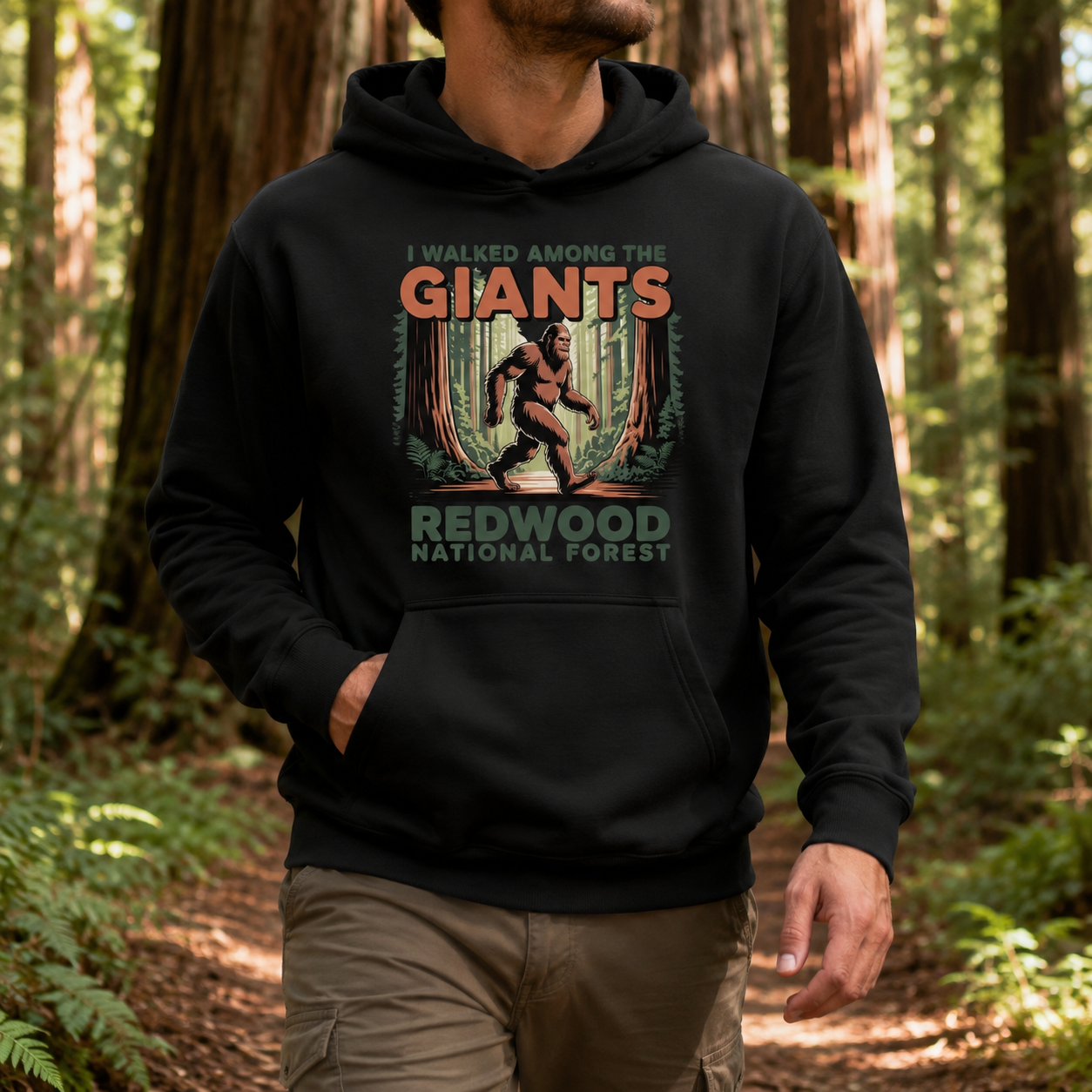 A man wearing a black hoodie with a vintage-style graphic that reads 'I WALKED AMONG THE GIANTS REDWOOD NATIONAL FOREST' and showing a Bigfoot figure, standing in a forest.