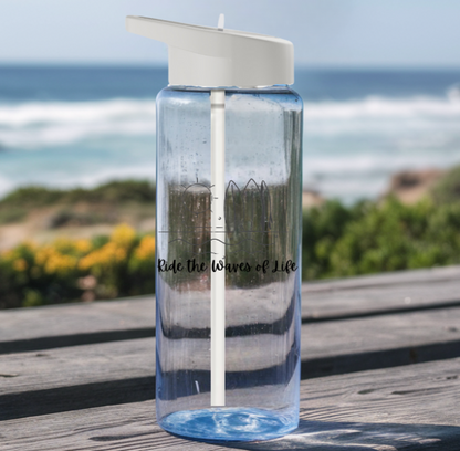 Clear water bottle with gray lid and clear straw with a picture of waves, surfboards, the sun and says Ride the Waves of Life sitting on a picnic table at the beach