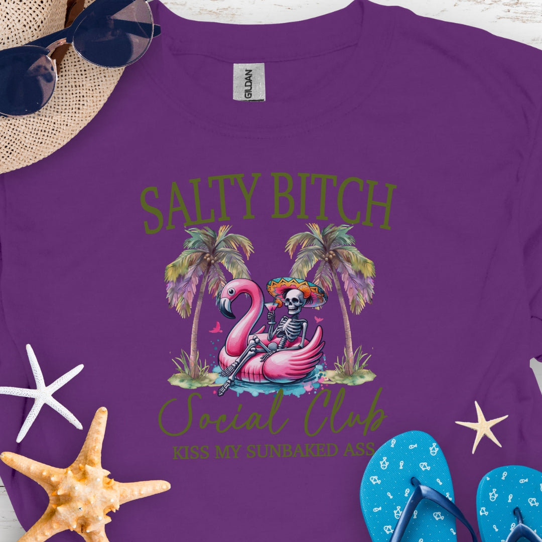 Purple t-shirt beach skeleton floating in flamingo floaty between two palm trees with text Salty Beach Social Club Kiss my sunkissed ass.