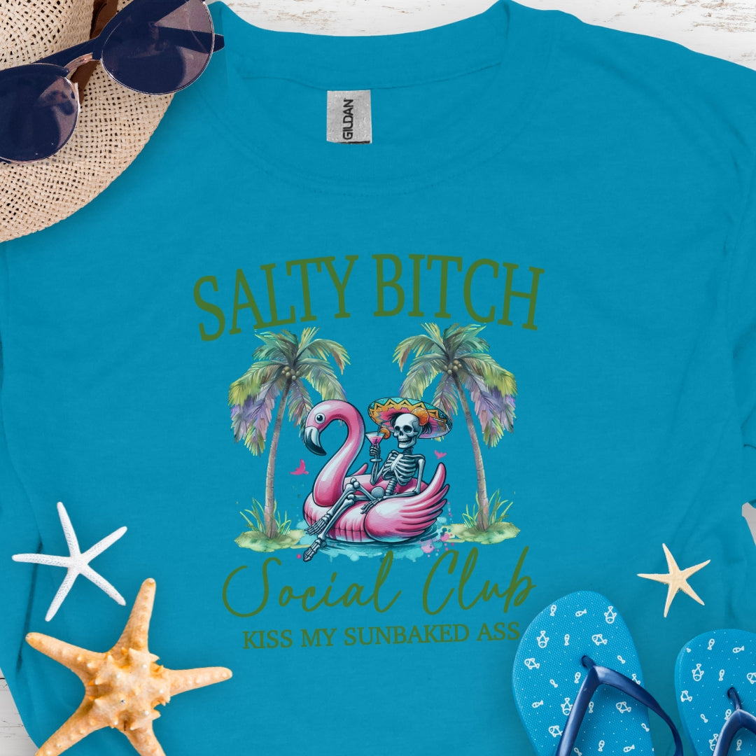 Tropical Blue t-shirt beach skeleton floating in flamingo floaty between two palm trees with text Salty Beach Social Club Kiss my sunkissed ass.