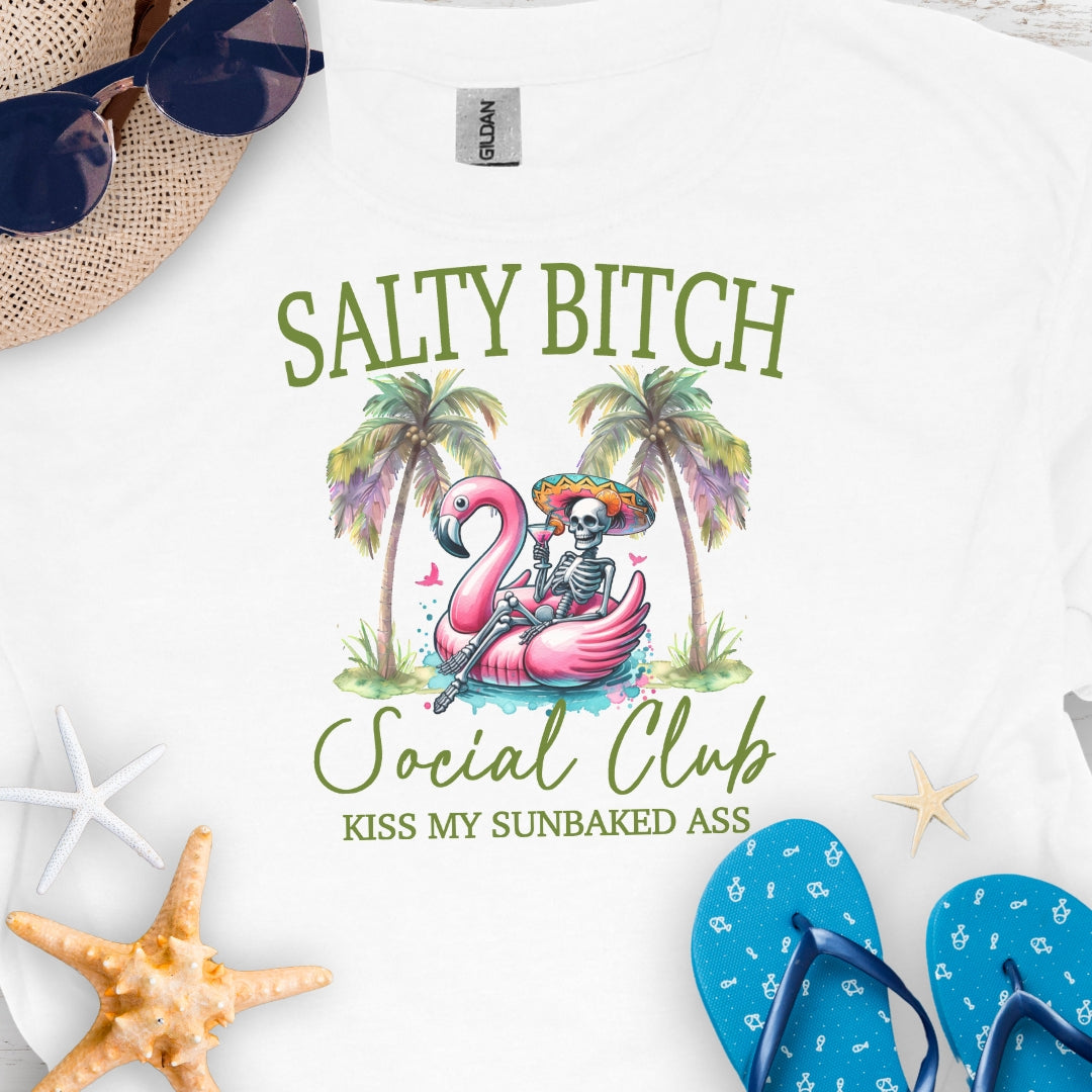 White t-shirt beach skeleton floating in flamingo floaty between two palm trees with text Salty Beach Social Club Kiss my sunkissed ass.