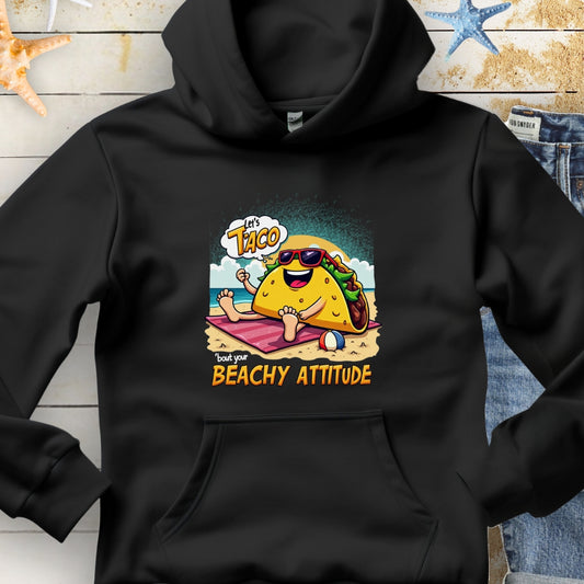 Black hoodie with a taco wearing sunglasses laying on a beach towel on the beach with the text Let's Taco 'bout your Beachy Attitude.