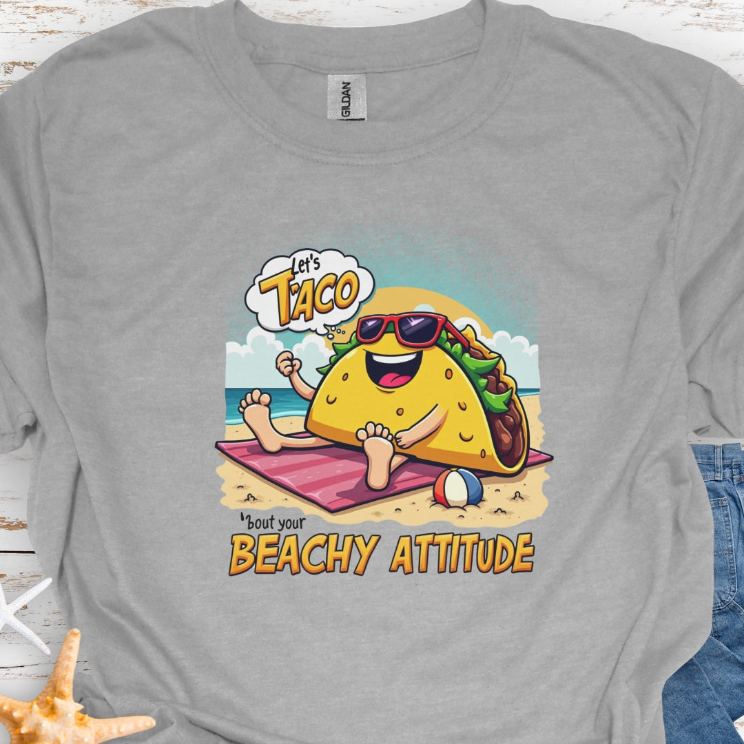 Gray  t-shirt with a taco wearing sunglasses laying on a beach towel on the beach with the text Let's Taco 'bout your Beachy Attitude.