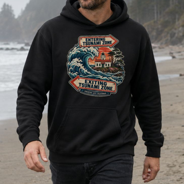 A man wearing a black pullover hoodie with a vintage-style tsunami warning graphic on the chest, standing on a sandy beach.