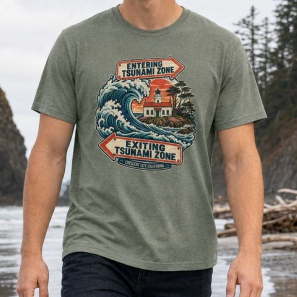 A man wearing a heathered military green t-shirt with a vintage-style design featuring a crashing ocean wave, coastal lighthouse, and vintage entering and exiting tsunami zone signs.