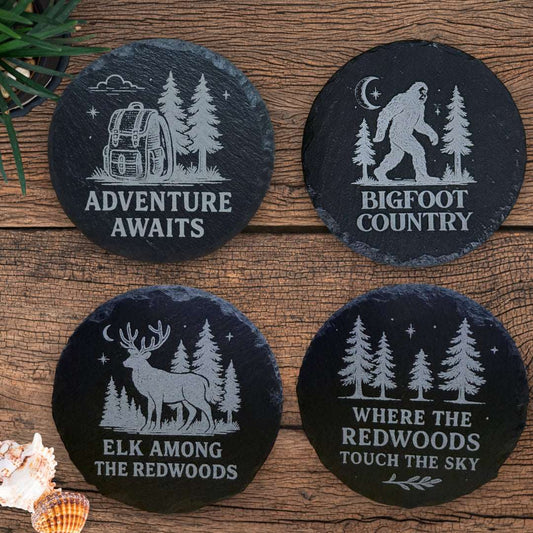 Four black slate coasters with nature-themed designs on a wooden surface.