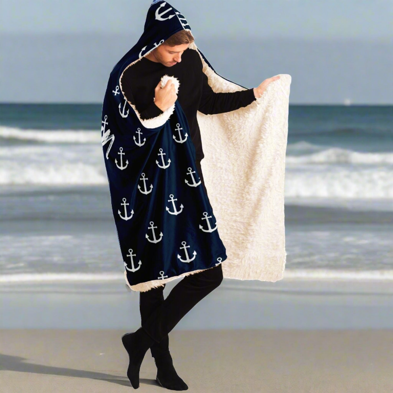 Person standing on the beach in a half open dark blue hooded blanket with white anchors and white writing that says The Ocean is my Anchor