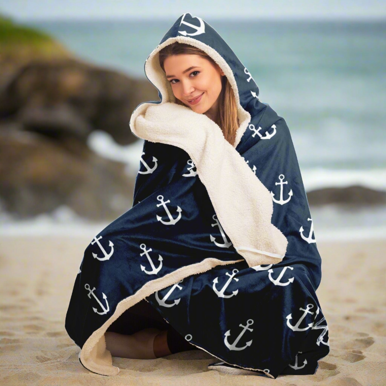 Person sitting on the beach wrapped in a dark blue hooded blanket with white anchors and white writing that says The Ocean is my Anchor