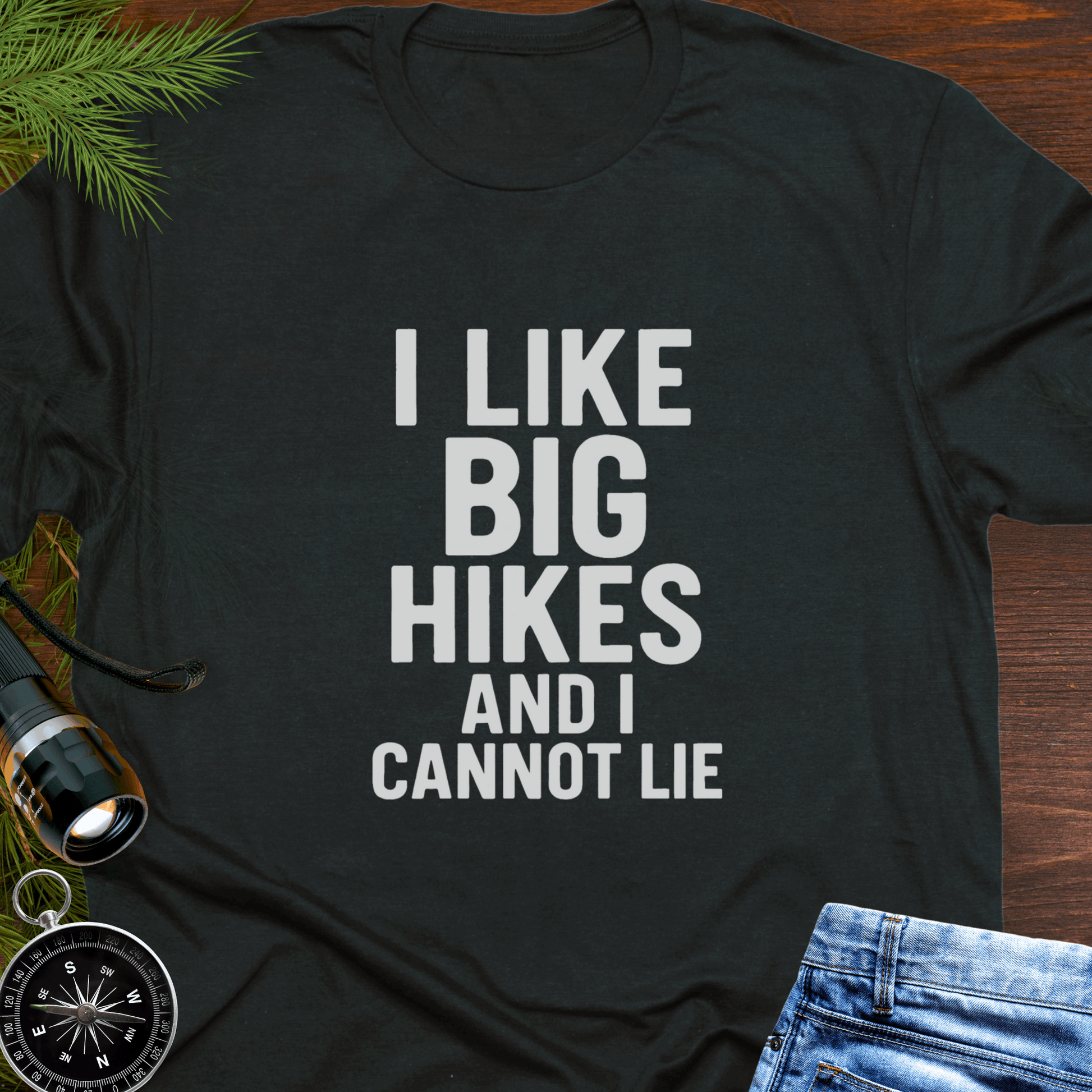 Black t-shirt with 'I LIKE BIG HIKES AND I CANNOT LIE' text on a wooden surface with hiking accessories.
