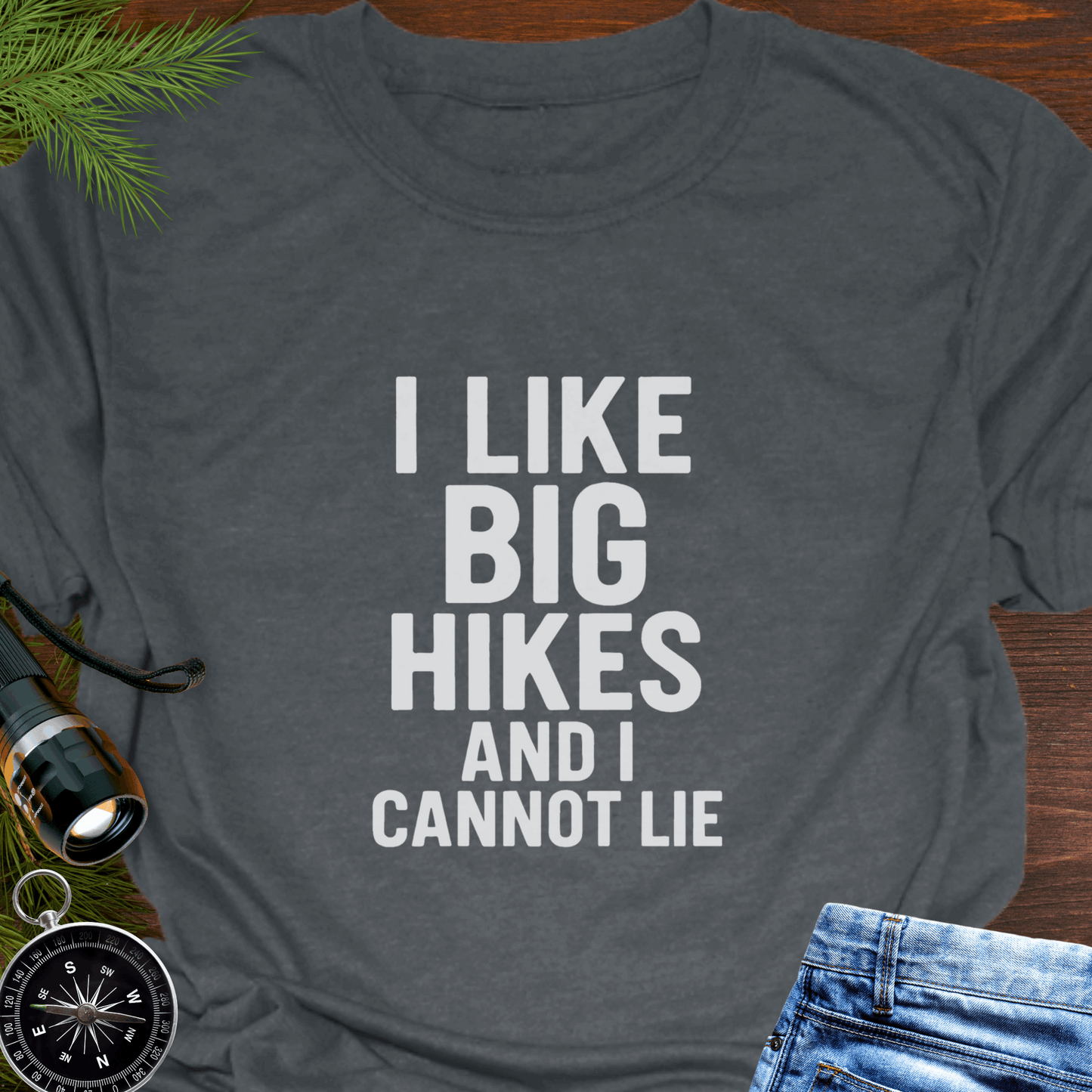 Gray t-shirt with 'I LIKE BIG HIKES AND I CANNOT LIE' text on a wooden surface with hiking accessories.