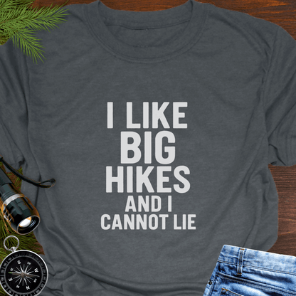 Gray t-shirt with 'I LIKE BIG HIKES AND I CANNOT LIE' text on a wooden surface with hiking accessories.