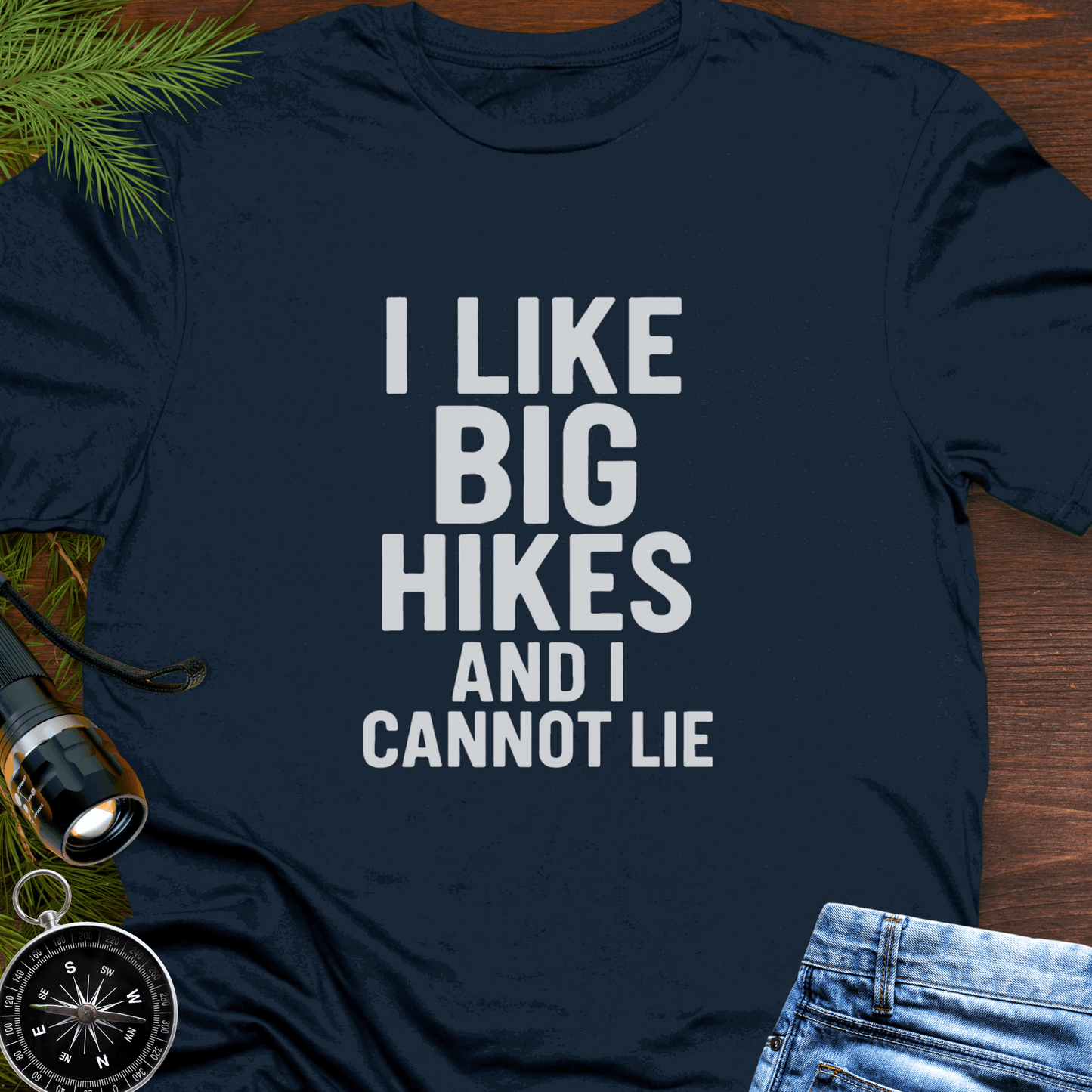Navy blue t-shirt with 'I LIKE BIG HIKES AND I CANNOT LIE' text on a wooden surface with hiking accessories.