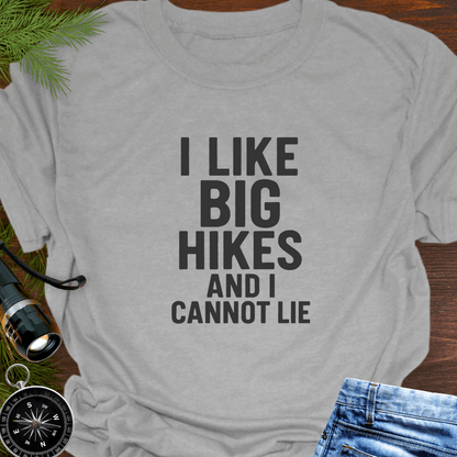 Gray t-shirt with 'I LIKE BIG HIKES AND I CANNOT LIE' text, surrounded by hiking accessories on a wooden surface.