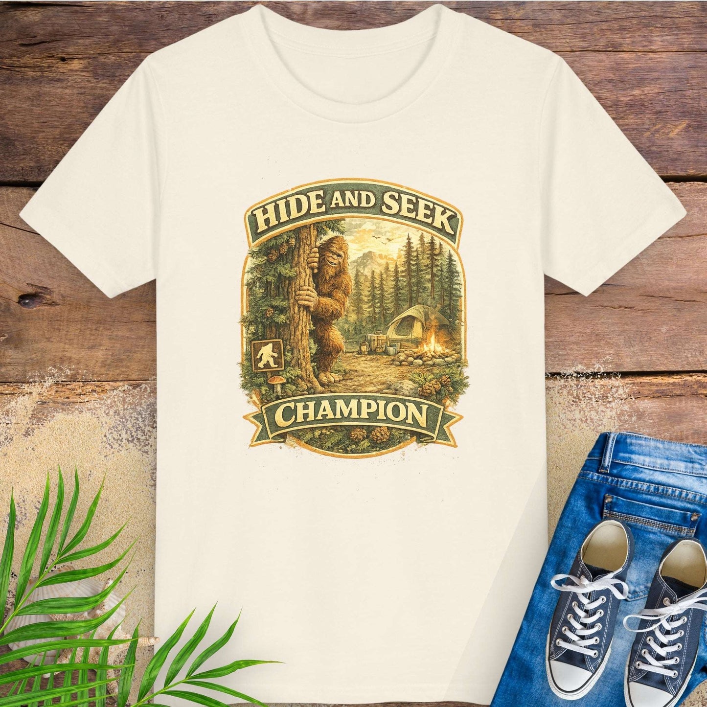 Light beige t-shirt with 'Hide and Seek Champion' graphic with Bigfoot peeking around a tree in a campsite.